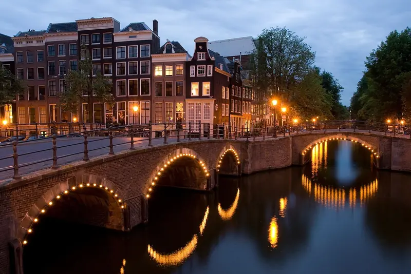 Pontes em arco iluminadas no cruzamento dos canais Keizersgracht e Reguliersgracht ao anoitecer, com casas estreitas de tijolo ao fundo