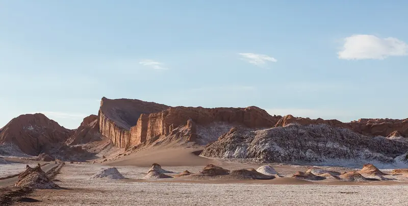 Paredão de rocha avermelhada do Anfiteatro no Valle de la Luna, com uma duna clara aos pés e o chão coberto de sal sob céu azul