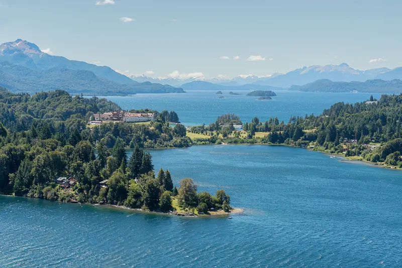 Península Llao Llao vista do alto, com bosques escuros, um grande edifício de telhado vermelho e o lago Nahuel Huapi azul até as montanhas nevadas
