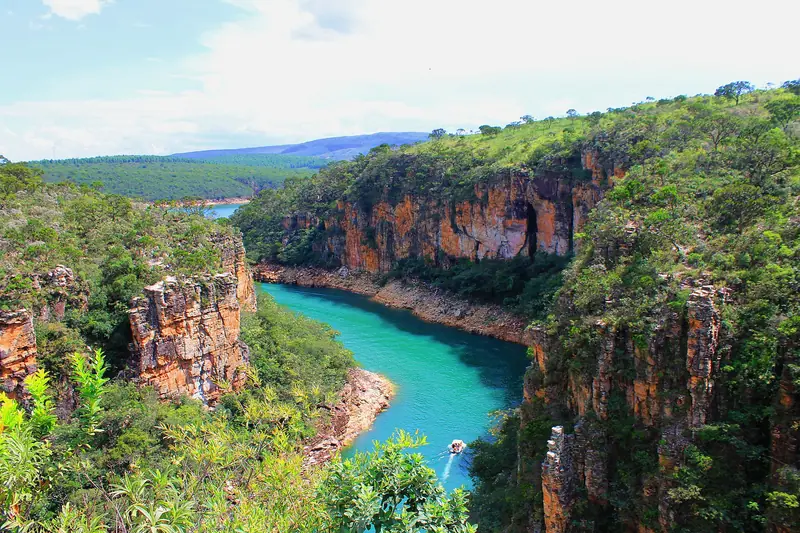 Vista do alto dos cânions de Capitólio: um braço de água verde-turquesa entre paredões alaranjados cobertos de mata, com uma lancha pequena lá embaixo