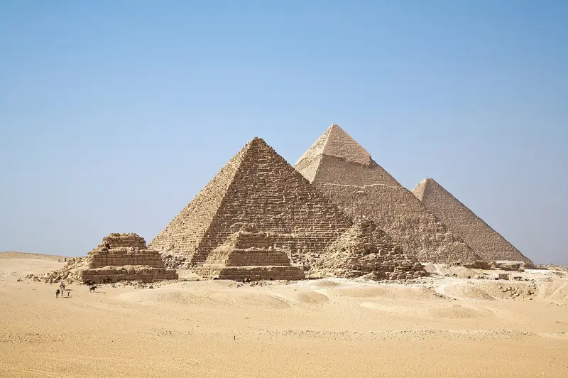 As três grandes pirâmides de Gizé sob céu azul limpo, com pirâmides menores das rainhas à frente e a areia clara do deserto em primeiro plano