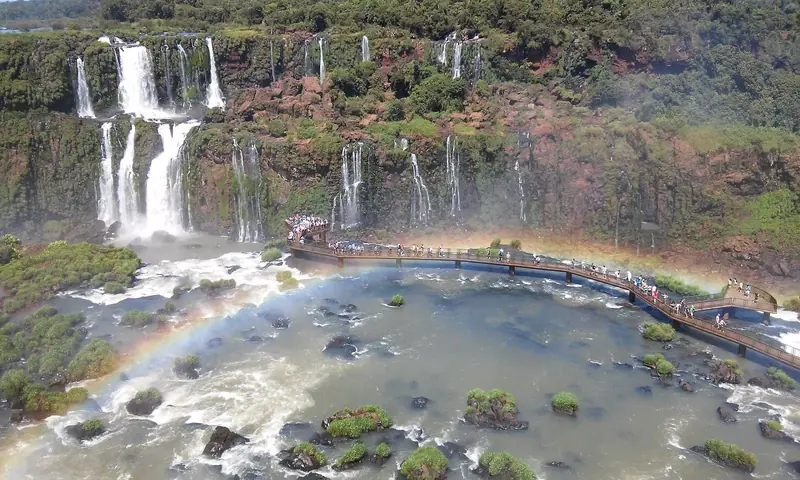 Passarela do lado brasileiro das Cataratas do Iguaçu sobre o rio, com quedas ao fundo e um arco-íris