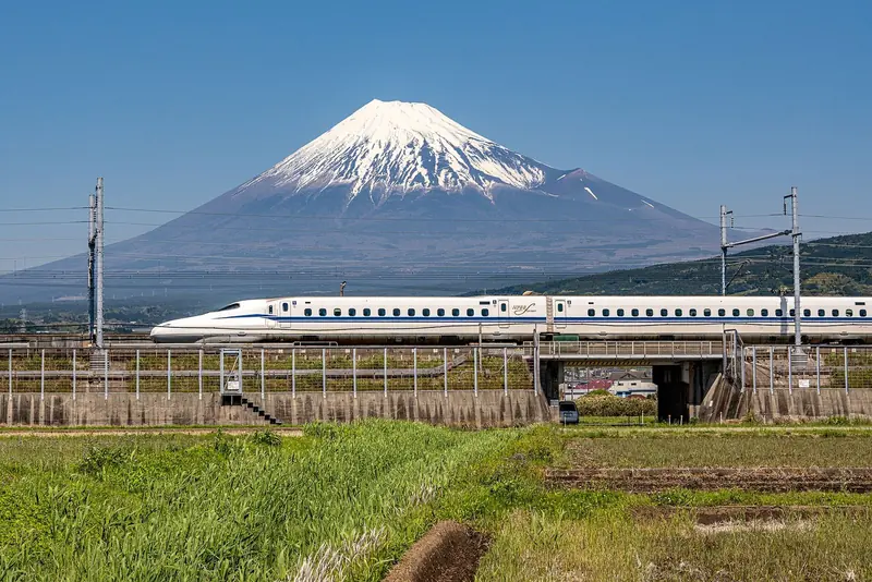 Trem-bala branco do Tokaido Shinkansen passando por um viaduto entre plantações, com o monte Fuji nevado ao fundo sob céu azul