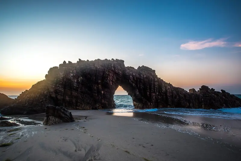 A Pedra Furada de Jericoacoara, arco de rocha escura na beira do mar, contra a luz do fim de tarde
