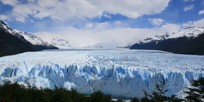 Frente do Glaciar Perito Moreno visto das passarelas, com o gelo azulado em pontas até o fundo do vale e montanhas nevadas dos dois lados