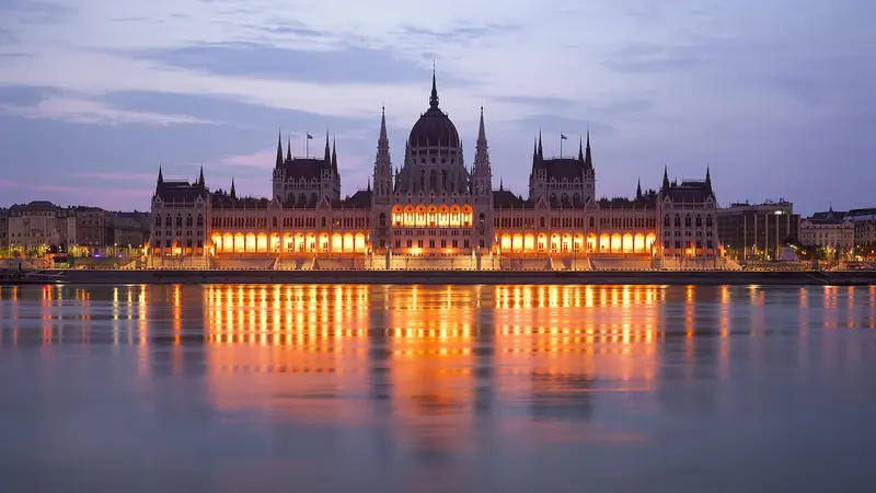 O Parlamento húngaro iluminado ao entardecer, refletido no Danúbio, com o domo central e as torres góticas sob céu lilás