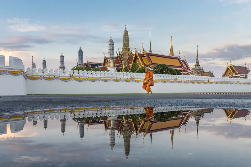 Muro branco do Grande Palácio de Bangkok com os telhados e as torres douradas do templo do Buda de Esmeralda atrás, um monge de túnica laranja caminhando e tudo refletido numa poça no chão