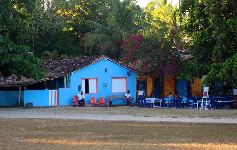Casa azul de janelas laranja e mesas de restaurante sob uma buganvília no Quadrado de Trancoso, com o gramado em primeiro plano